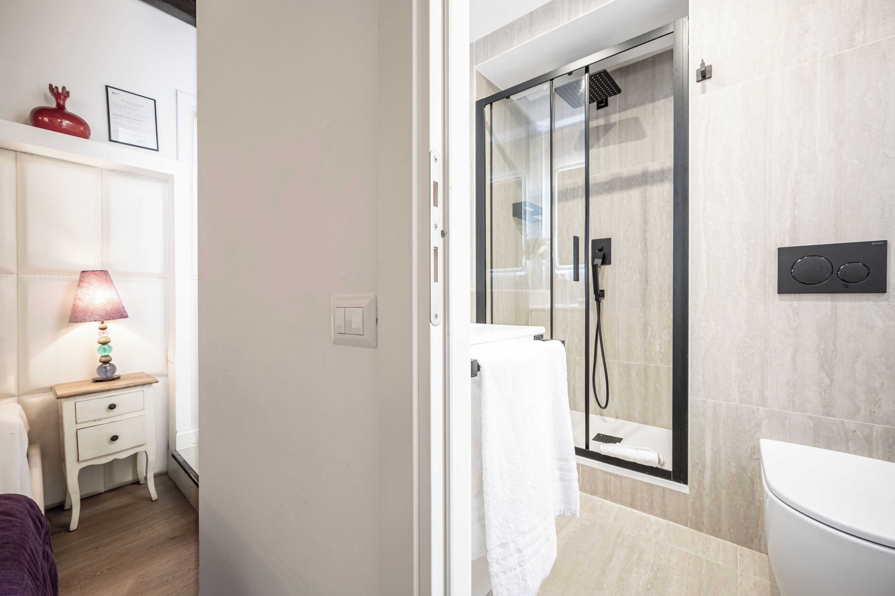A bathroom with a glass-enclosed shower, a white toilet, and a bedside table with a lamp.