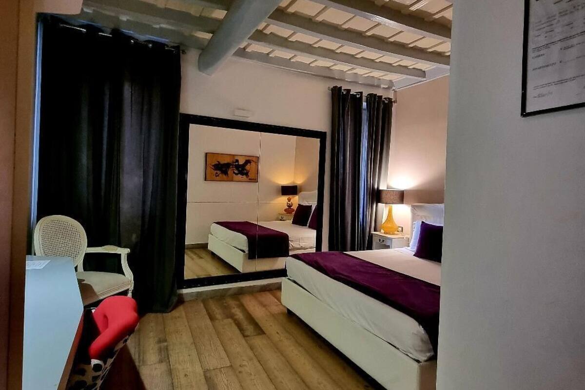 A compact hotel room with a wooden floor, a white bed with a purple blanket, a small desk with a chair, and a nightstand with a lamp.