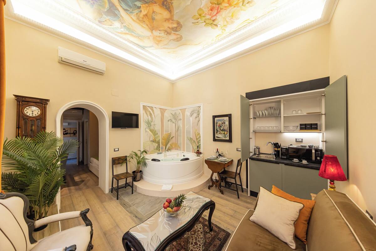 A spacious living area with a large window, a seating area with a sofa and armchairs, a coffee table with fruit, a hot tub, and a kitchen area with a microwave and sink.