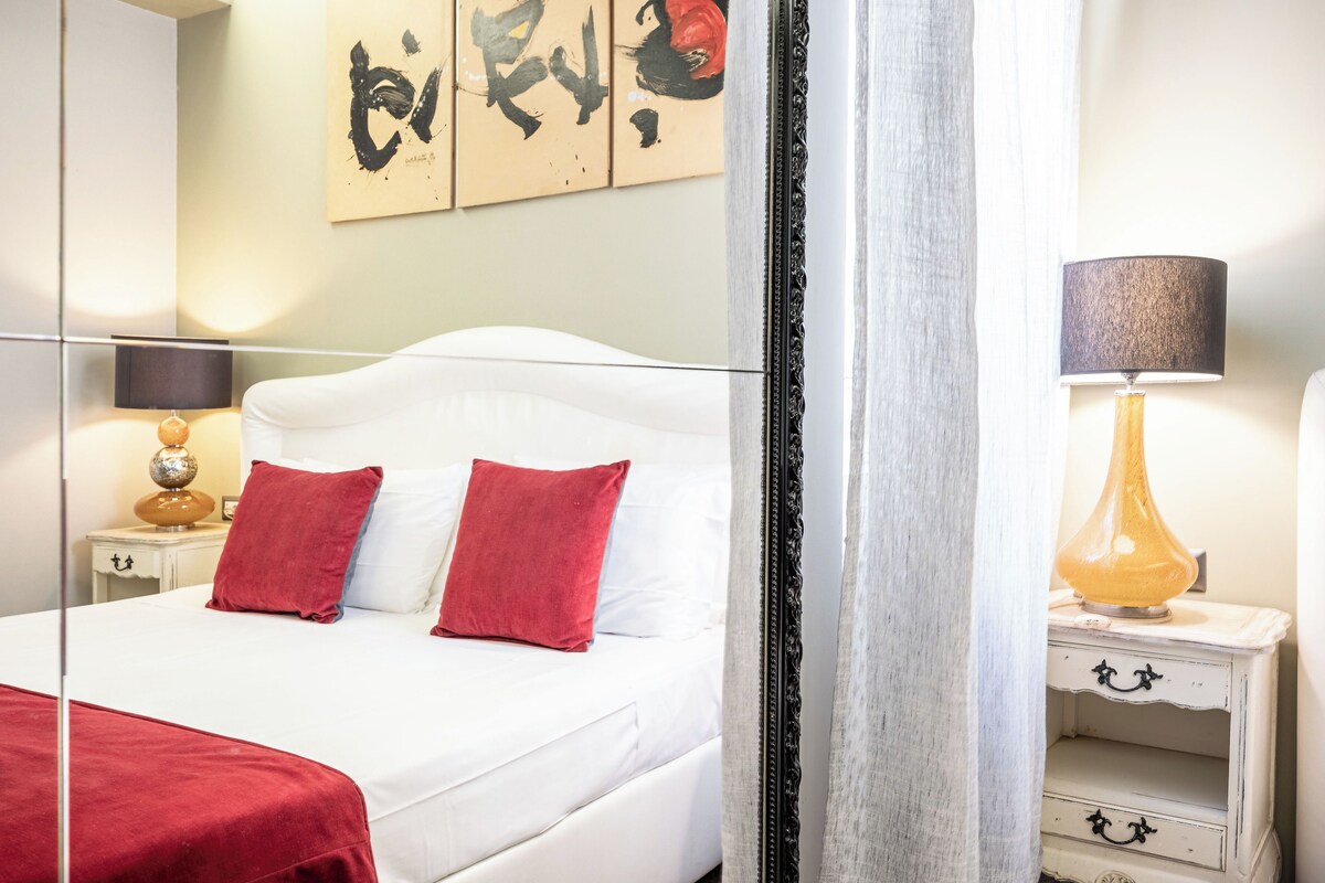 A hotel room with a white bed, red pillows, a nightstand with a lamp, and a wall with abstract art.