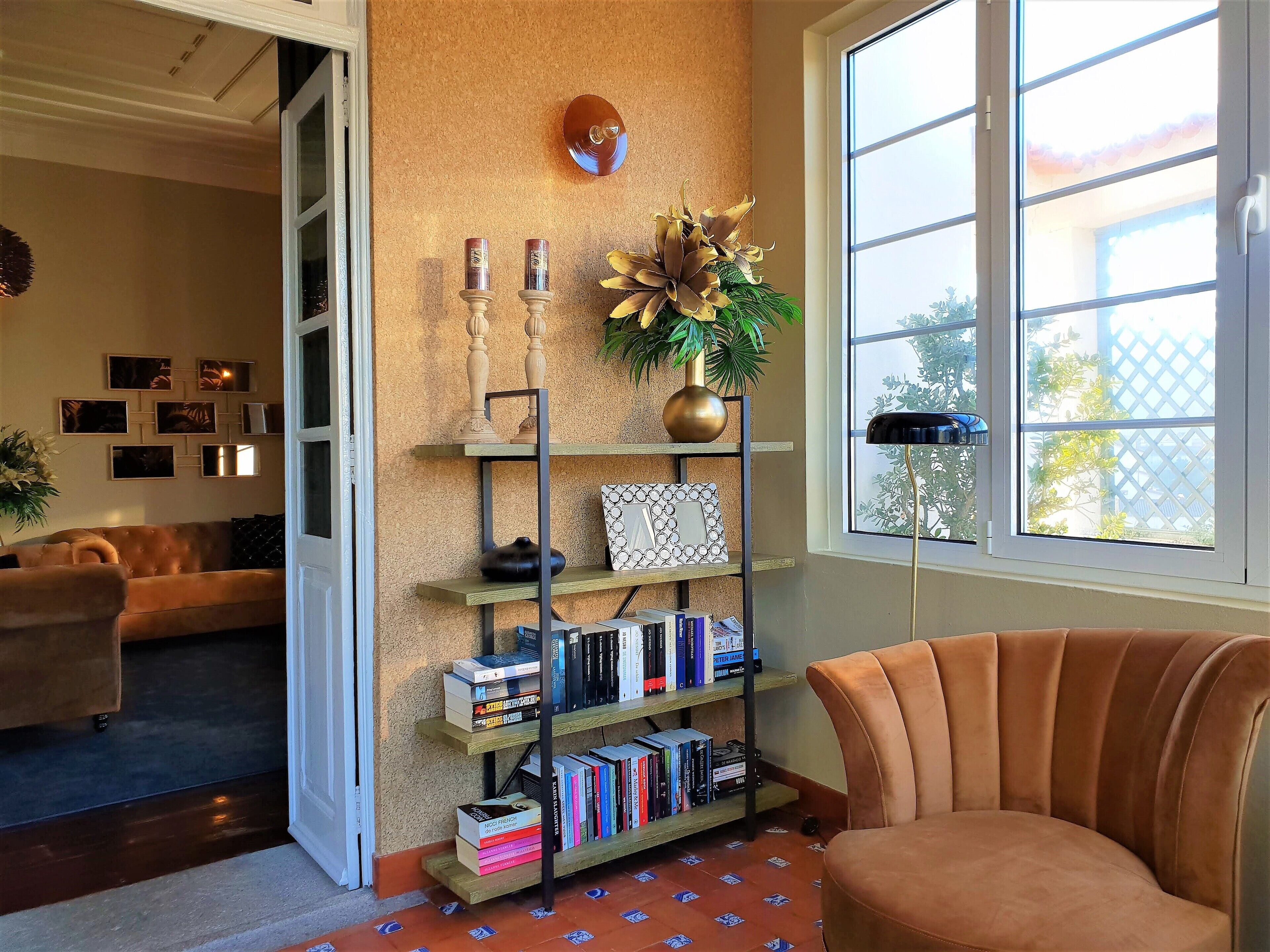 A cozy living room with a brown sofa, a bookshelf filled with books, a decorative plant, and a large window offering a view of the outside.