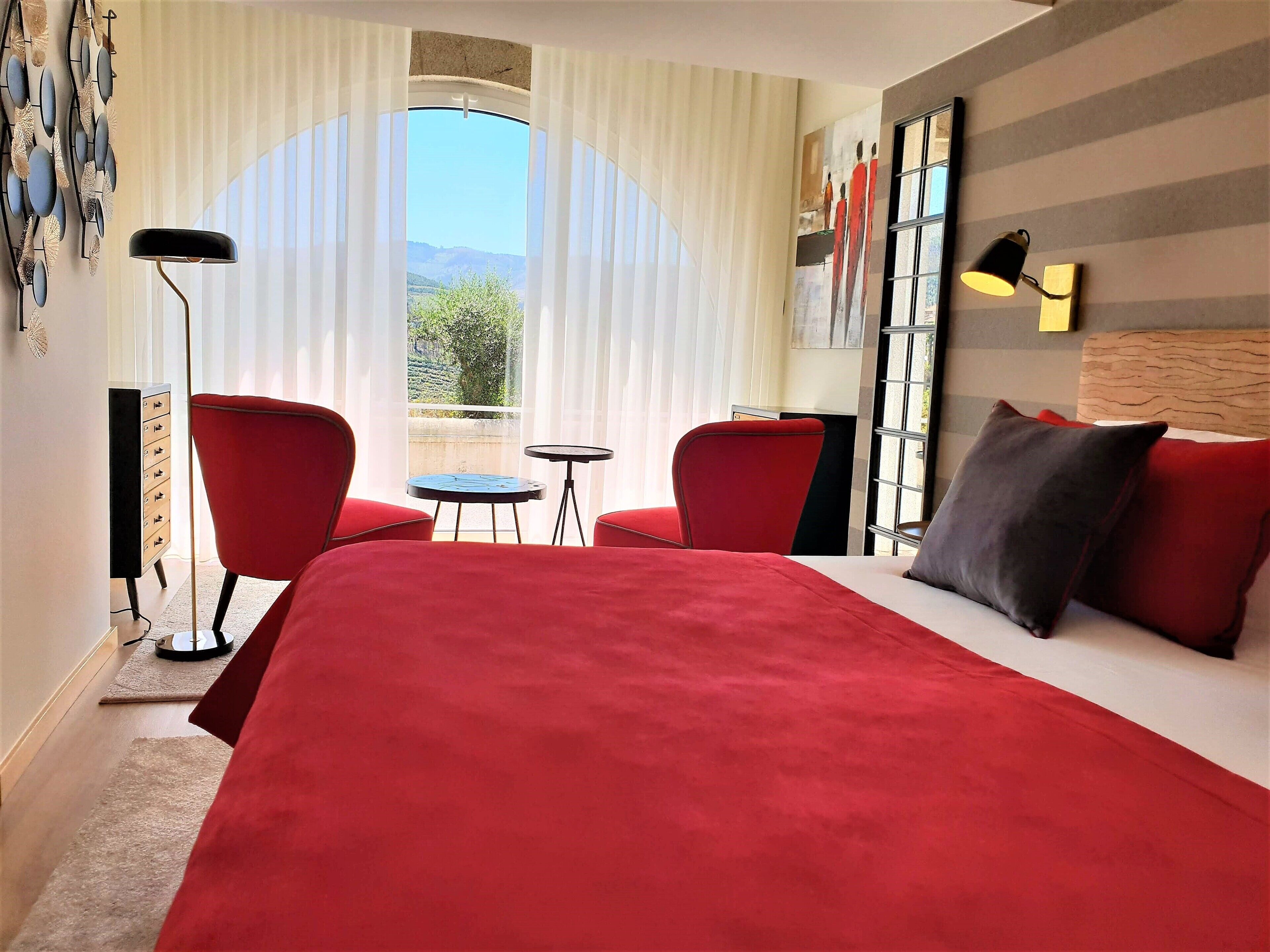 A hotel room with a large bed, two red chairs, a small table, a floor lamp, and a view of greenery through the window.