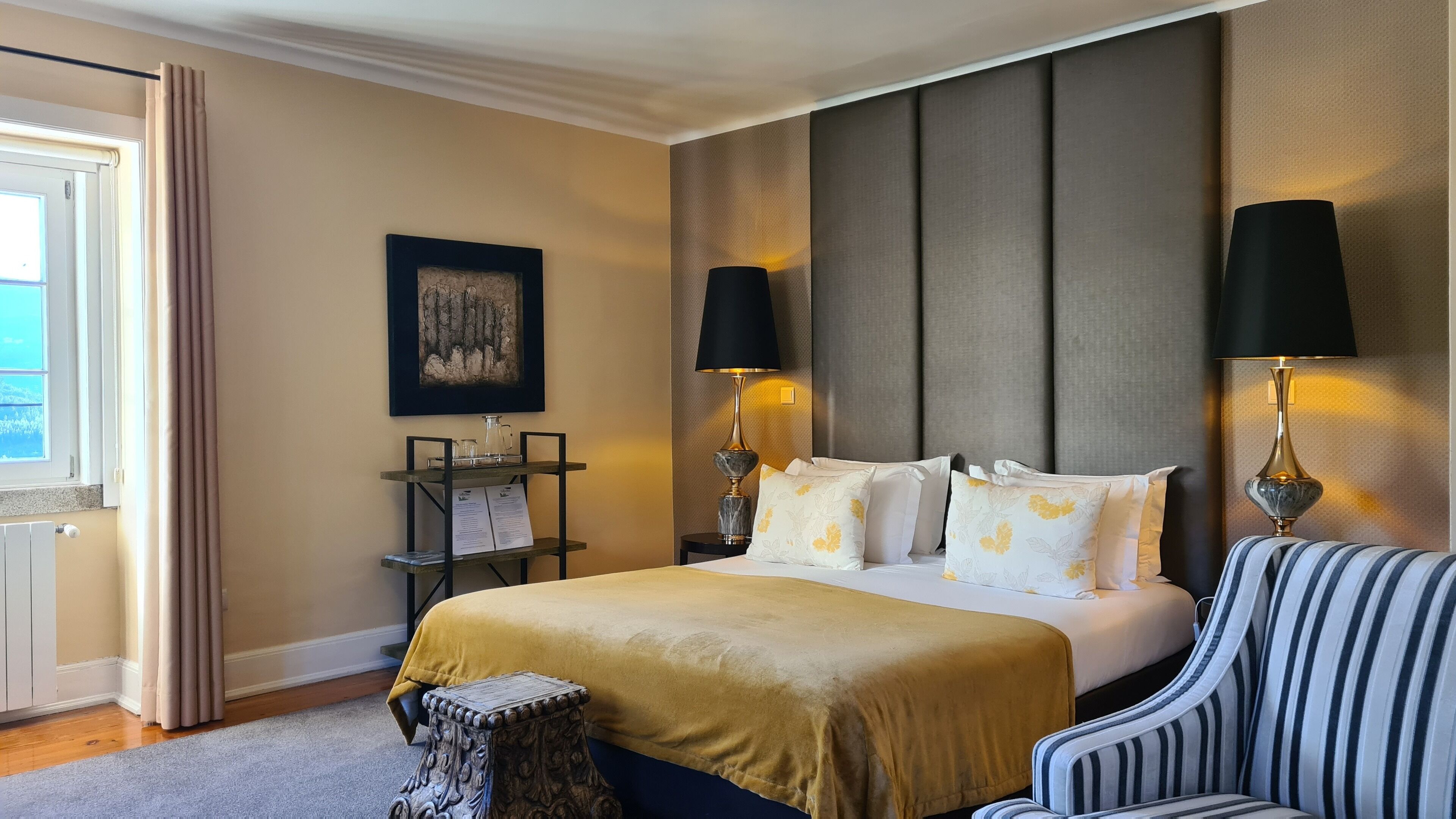 A hotel room with a bed, two bedside lamps, a small side table, a chair, a picture on the wall, and a window with curtains.