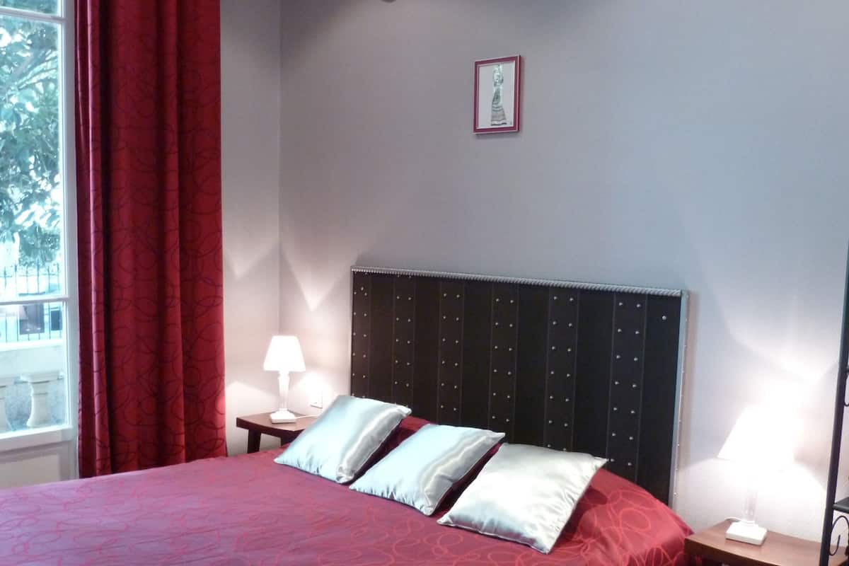 Un lit bien fait, recouvert d’une couvre-lit à motifs rouges, deux tables de chevet avec des lampes et un tableau encadré au mur.