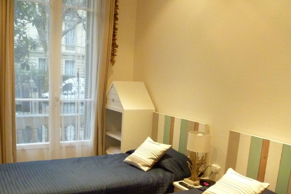 Une chambre d’hôtel avec deux lits, une fenêtre avec des rideaux, une table de chevet avec une lampe et une petite étagère.