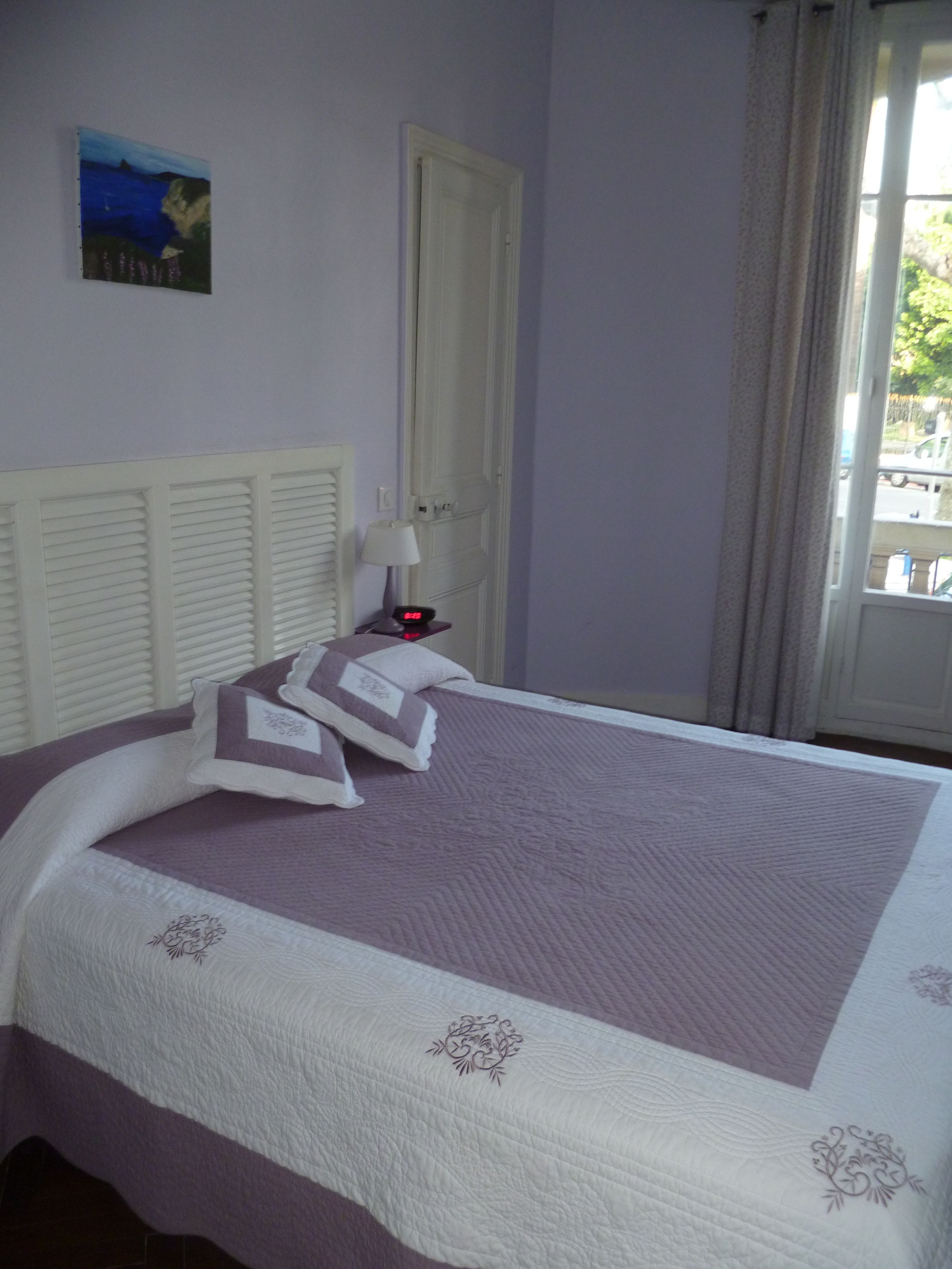 Uma cama bem arrumada com colcha roxa e branca, cabeceira branca e um quadro pendurado na parede.