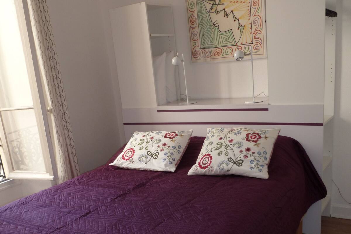 Une chambre à coucher avec un lit, une literie violette, deux coussins décoratifs, un miroir fixé au mur et une fenêtre avec des rideaux.