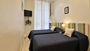 Habitación de hotel con dos camas, un televisor de pantalla plana montado en la pared, una ventana con cortinas y una mesita con lámpara.
