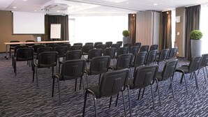 Un salón de conferencias con filas de sillas, una pantalla de proyector, una pizarra blanca y un amplio ventanal con cortinas.