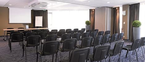 Une salle de conférence avec des rangées de chaises, un écran de projection, un tableau blanc et une grande fenêtre avec des rideaux.