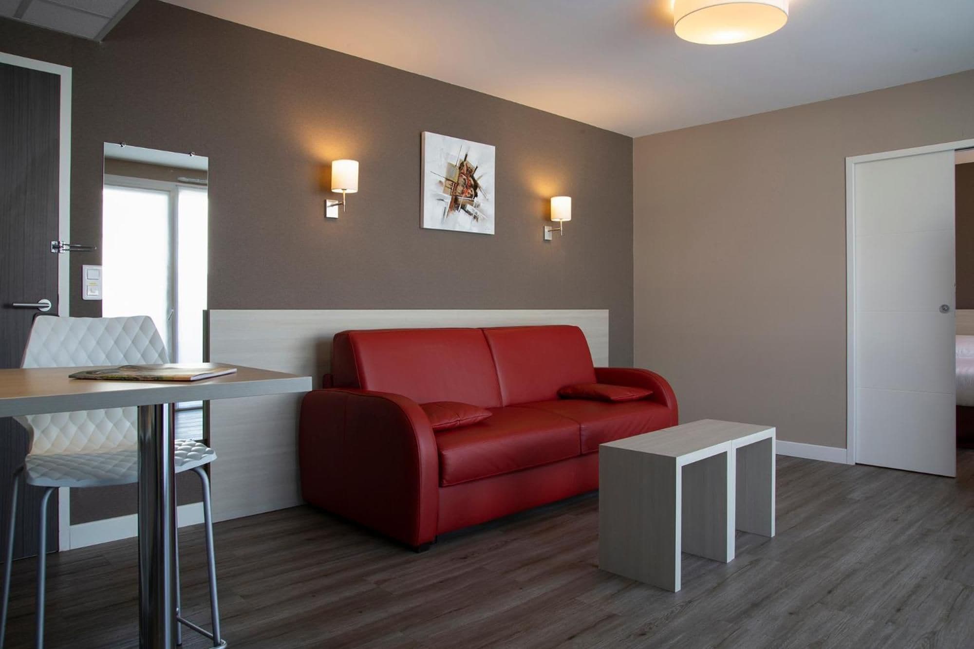 A modern room with a red sofa, a white table, and a small white side table. There are two wall-mounted lights and a framed picture on the wall.