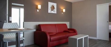 A modern room with a red sofa, a white table, and a small white side table. There are two wall-mounted lights and a framed picture on the wall.