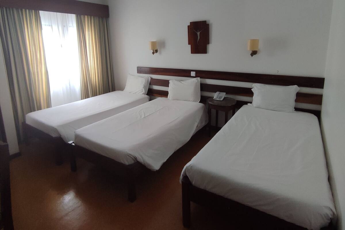 Une chambre d’hôtel avec trois lits simples, une tête de lit en bois, une table de chevet avec un téléphone et une horloge murale.