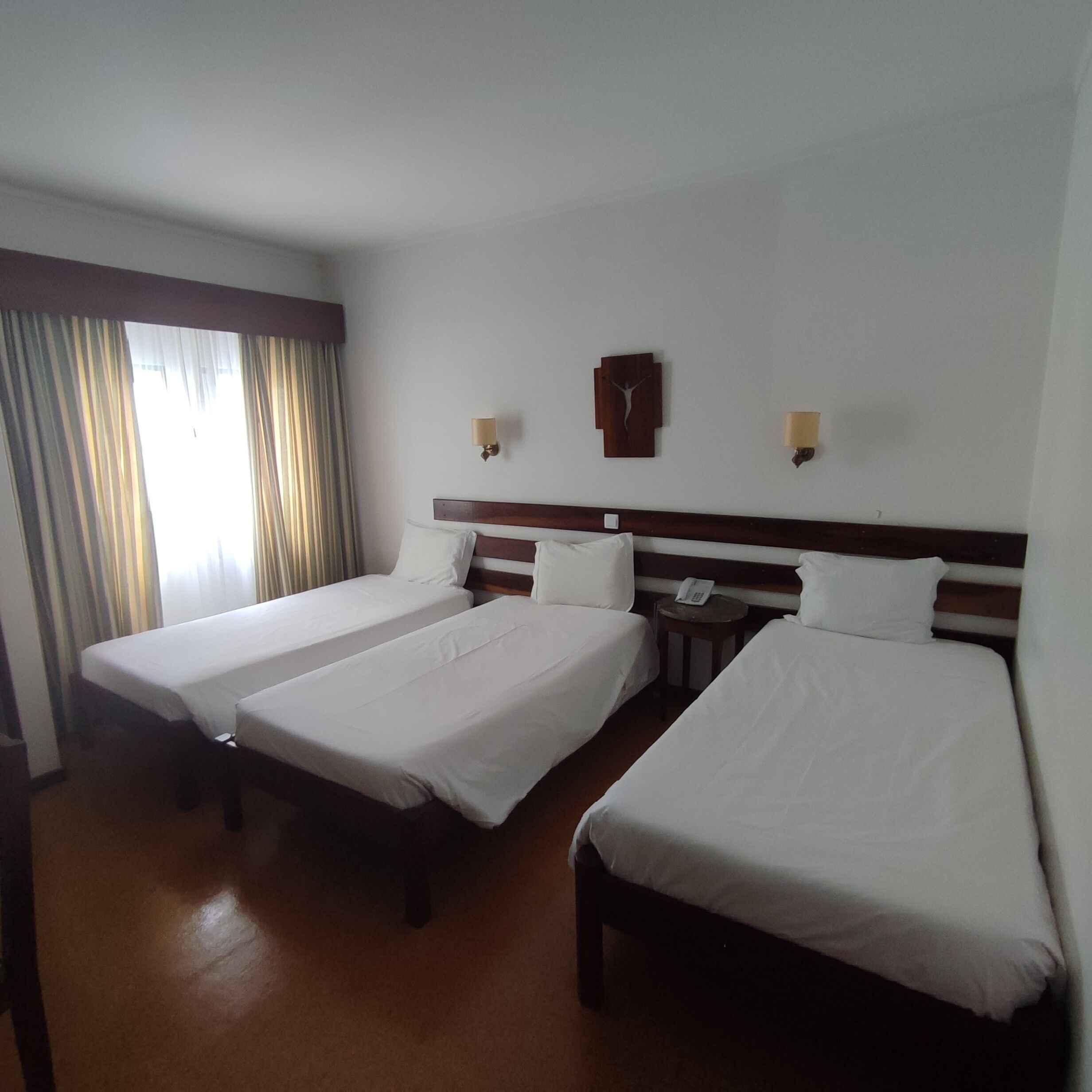 Une chambre d’hôtel avec trois lits simples, une tête de lit en bois, une table de chevet avec un téléphone et une horloge murale.