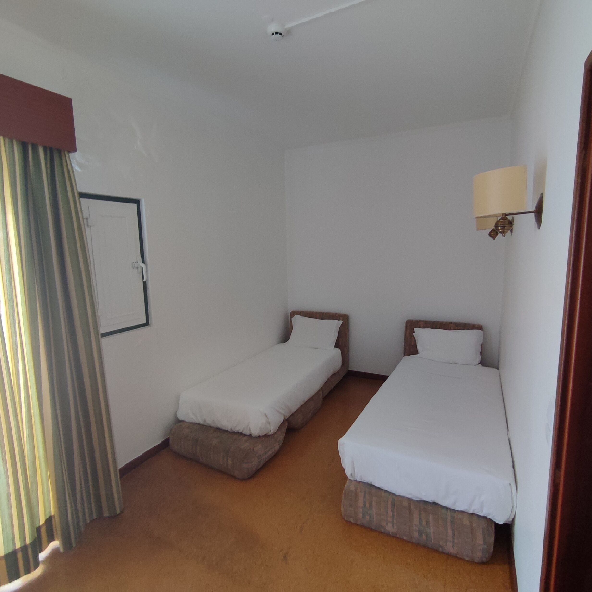 Ein Hotelzimmer mit zwei Einzelbetten, einem kleinen Tisch, einer Lampe und einem Vorhang.
