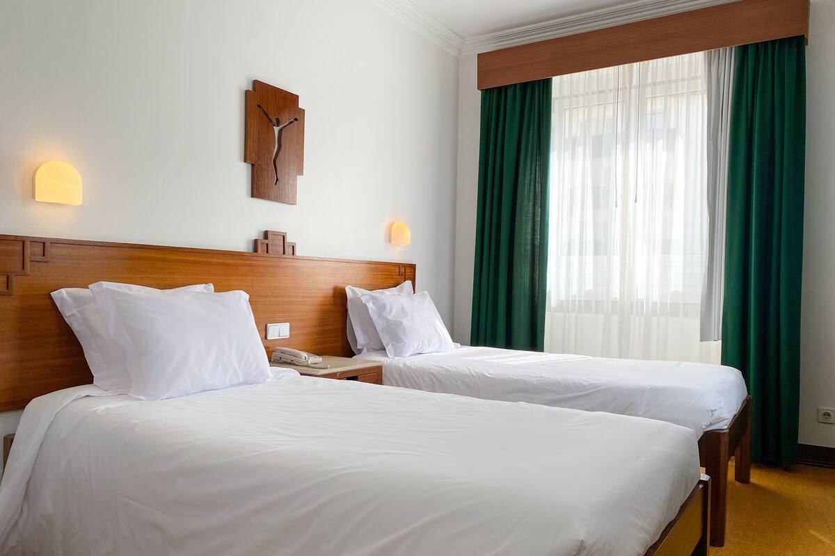 Une chambre d’hôtel avec deux lits, une tête de lit en bois, une horloge murale et une fenêtre avec des rideaux verts.