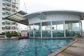 Un área de piscina moderna con una estructura de cristal, una piscina cristalina y una vista de un edificio de gran altura al fondo.