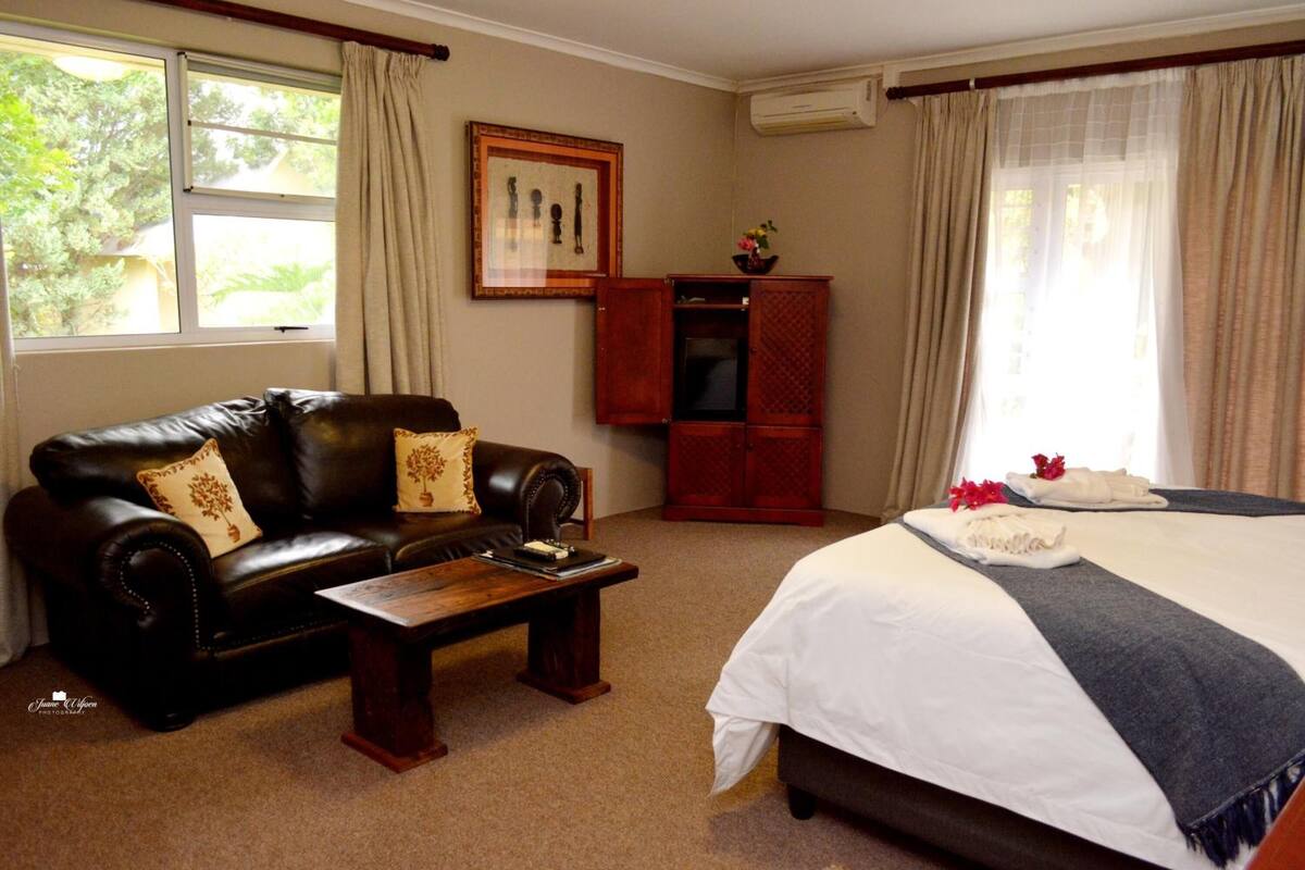 A hotel room with a leather sofa, a wooden coffee table, a bed with white bedding and a blue throw, a TV cabinet, and a framed picture on the wall.