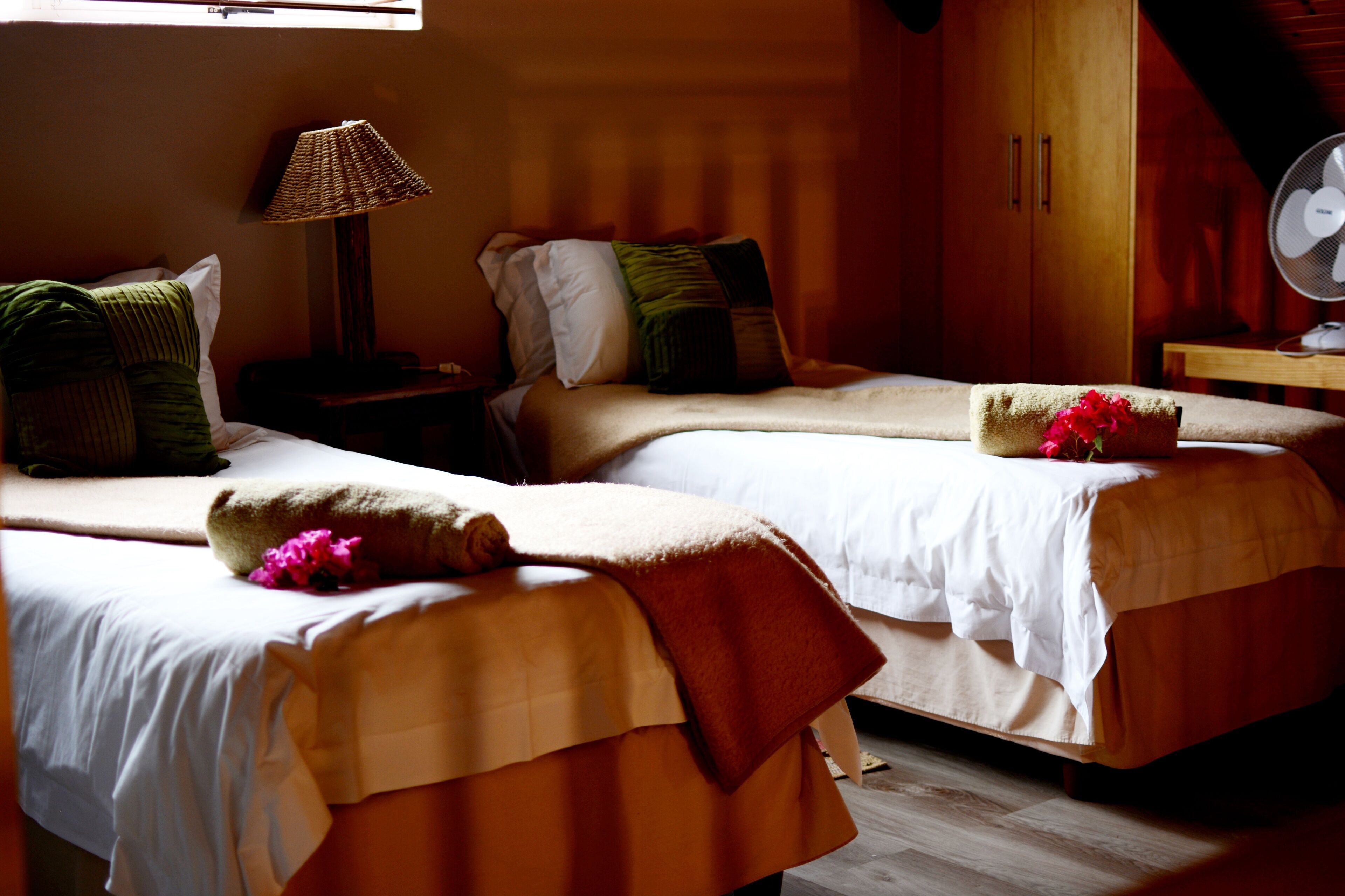 A hotel room with two beds, a bedside table with a lamp, a wooden wardrobe, and a fan.