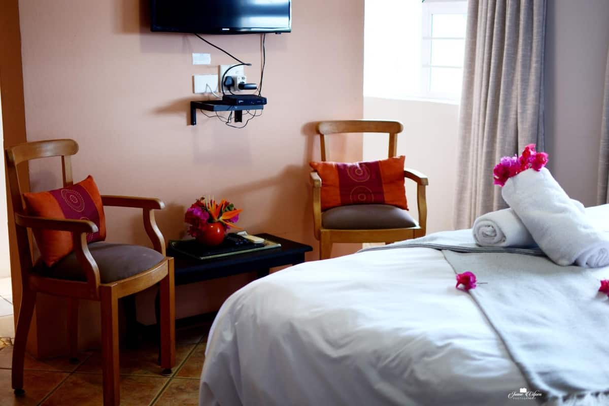 A hotel room with a bed, two wooden chairs, a small table with a vase of flowers, and a flat-screen TV mounted on the wall.
