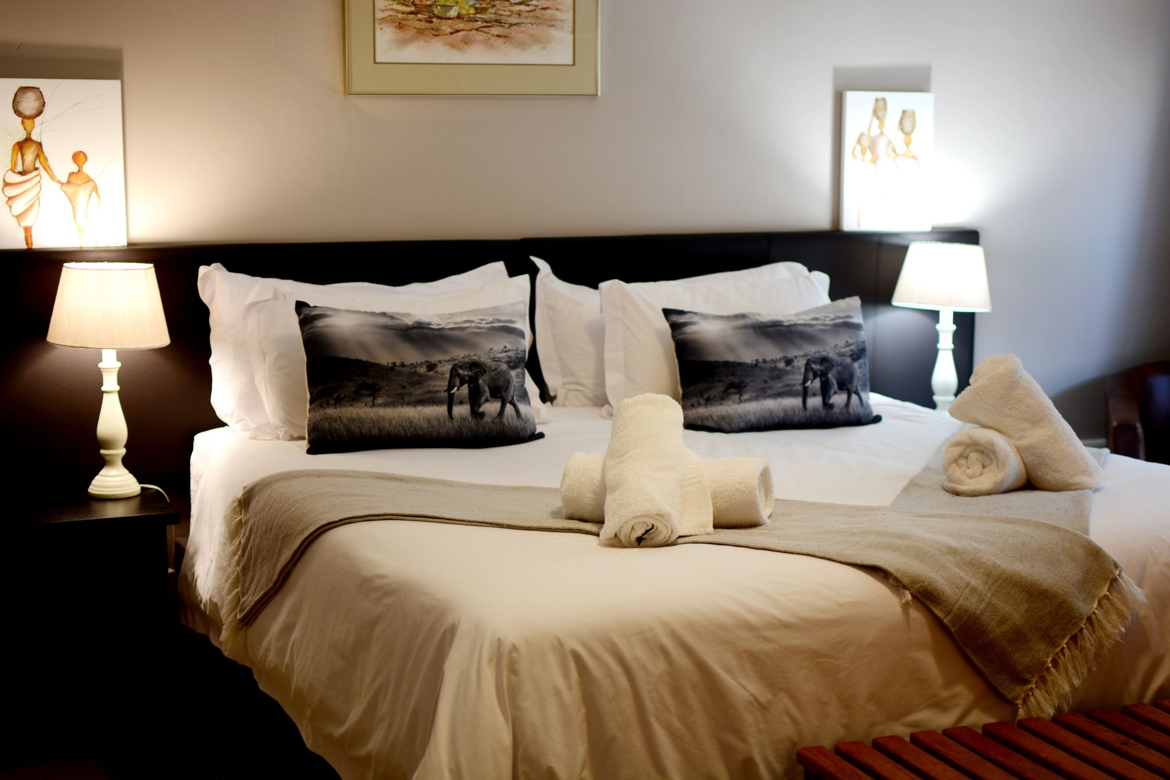Una cama bien hecha con sábanas blancas, dos almohadas con diseño de paisaje blanco y negro, y un par de toallas enrolladas.