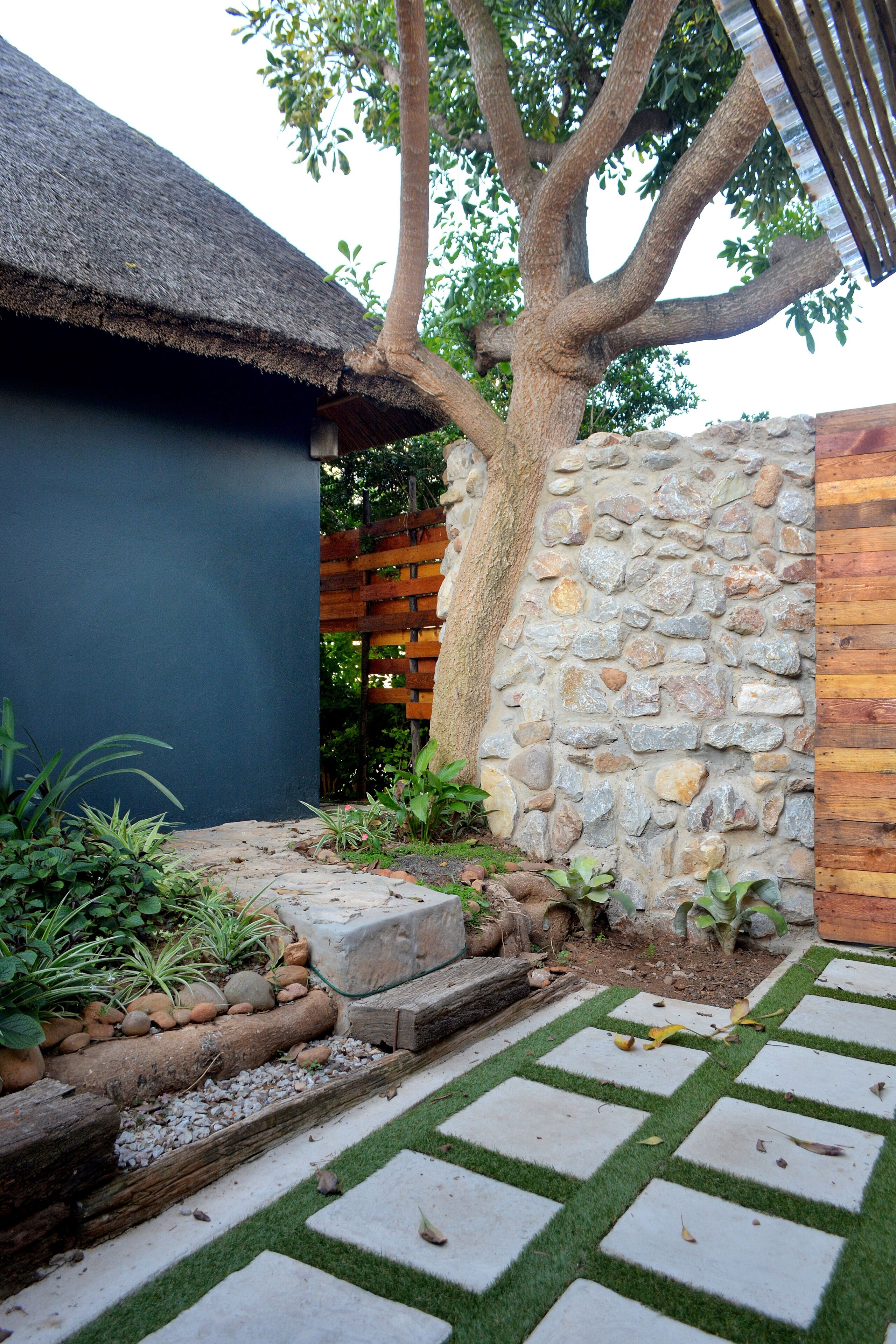 A stone wall with a wooden fence, a thatched-roof structure, and a paved walkway with grass inlays.