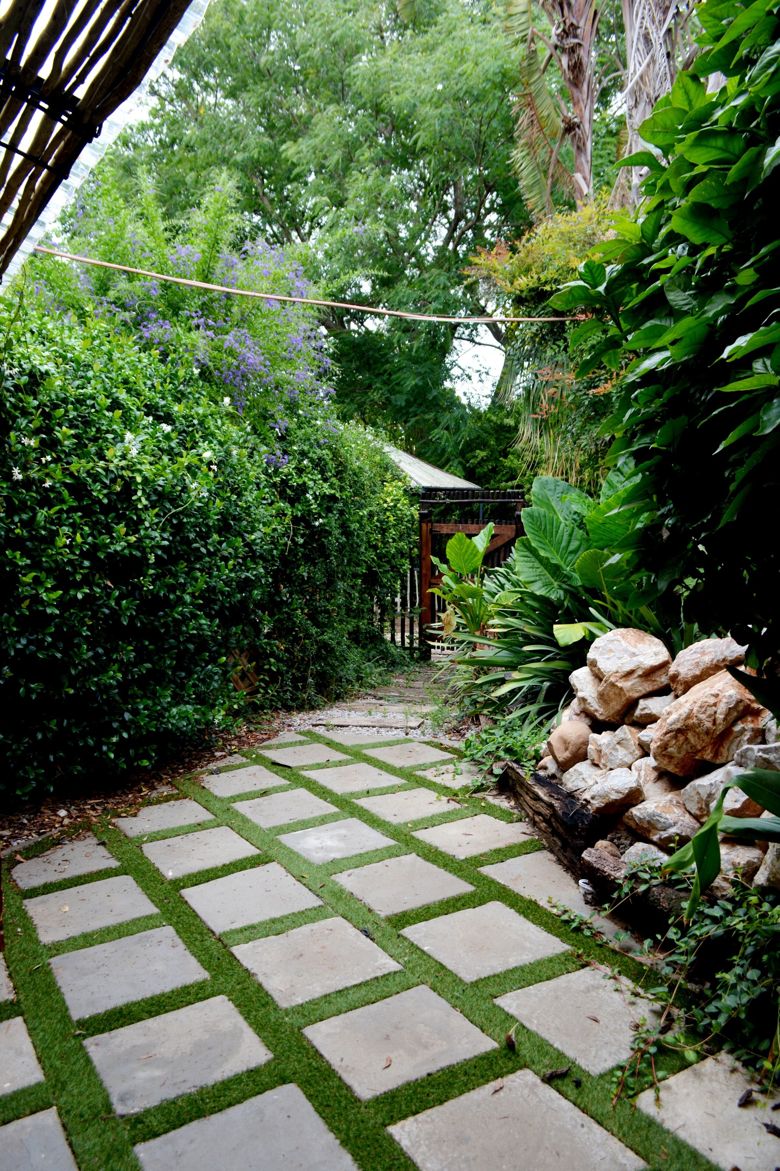 A stone pathway leading to a wooden structure surrounded by lush greenery and large rocks.