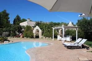 A swimming pool with lounge chairs and a pergola, surrounded by greenery and a stone building.