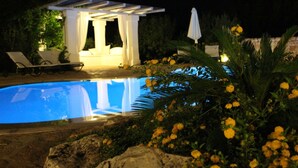 Un área de piscina nocturna con una zona de estar cubierta y sillones reclinables.