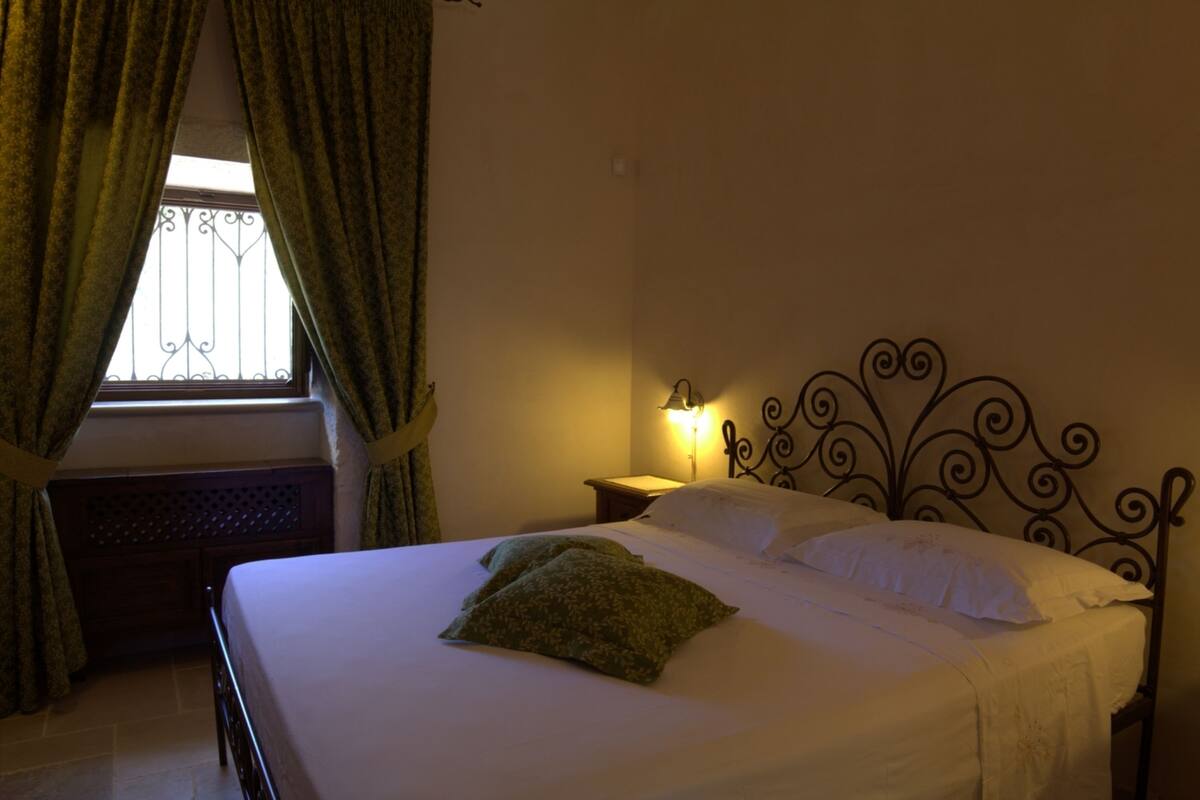 A neatly made bed with white linens and a green pillow, a bedside table with a lamp, and a window with curtains.