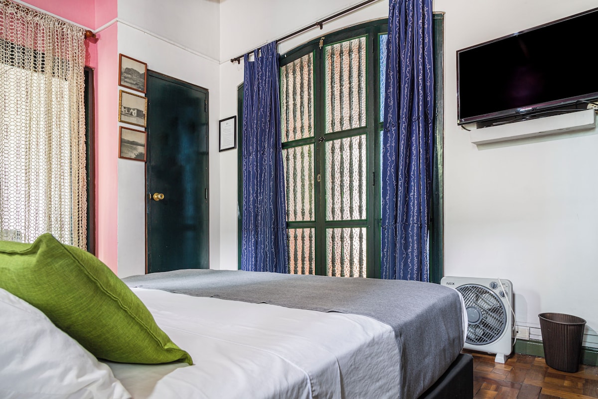 A bedroom with a bed, a wall-mounted TV, a window with curtains, and a standing fan.