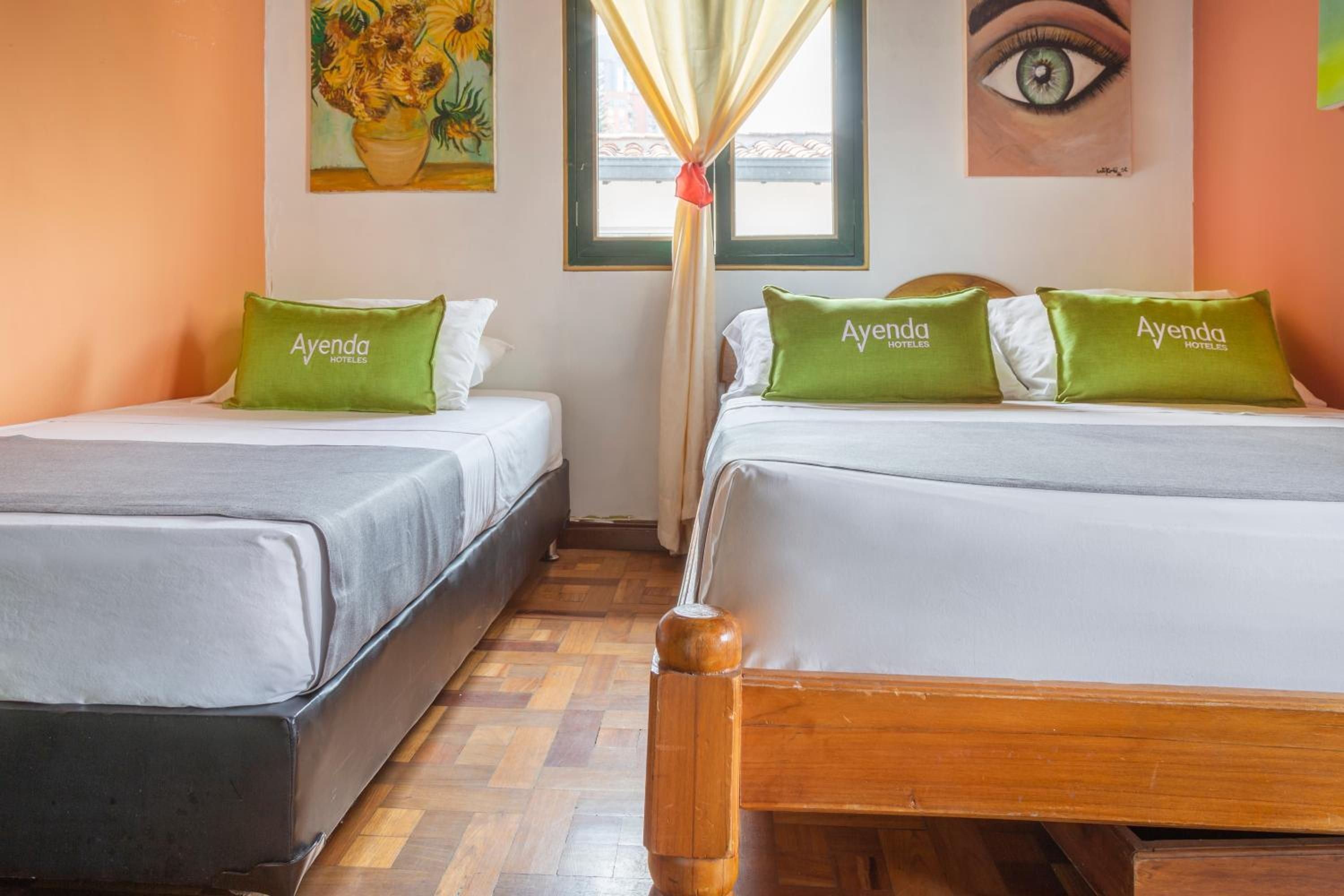 A hotel room with two beds, each with a green pillow labeled 'Ayenda Hoteles,' a painting of sunflowers, and a window with curtains.