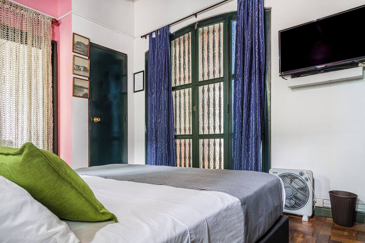 Une chambre à coucher avec un lit, un téléviseur fixé au mur, une fenêtre avec des rideaux et un ventilateur sur pied.