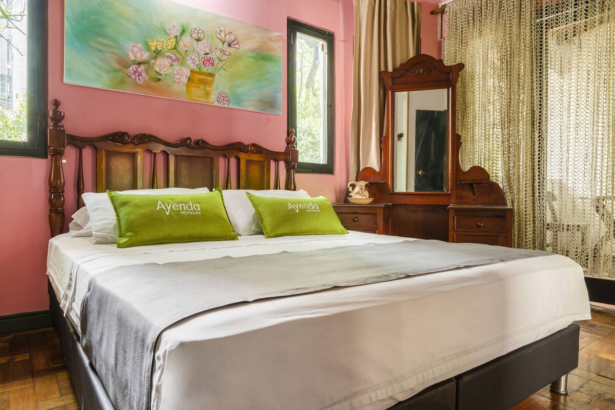 Une chambre d’hôtel avec un lit, une tête de lit en bois, un tableau de fleurs, un miroir et une fenêtre avec des rideaux.