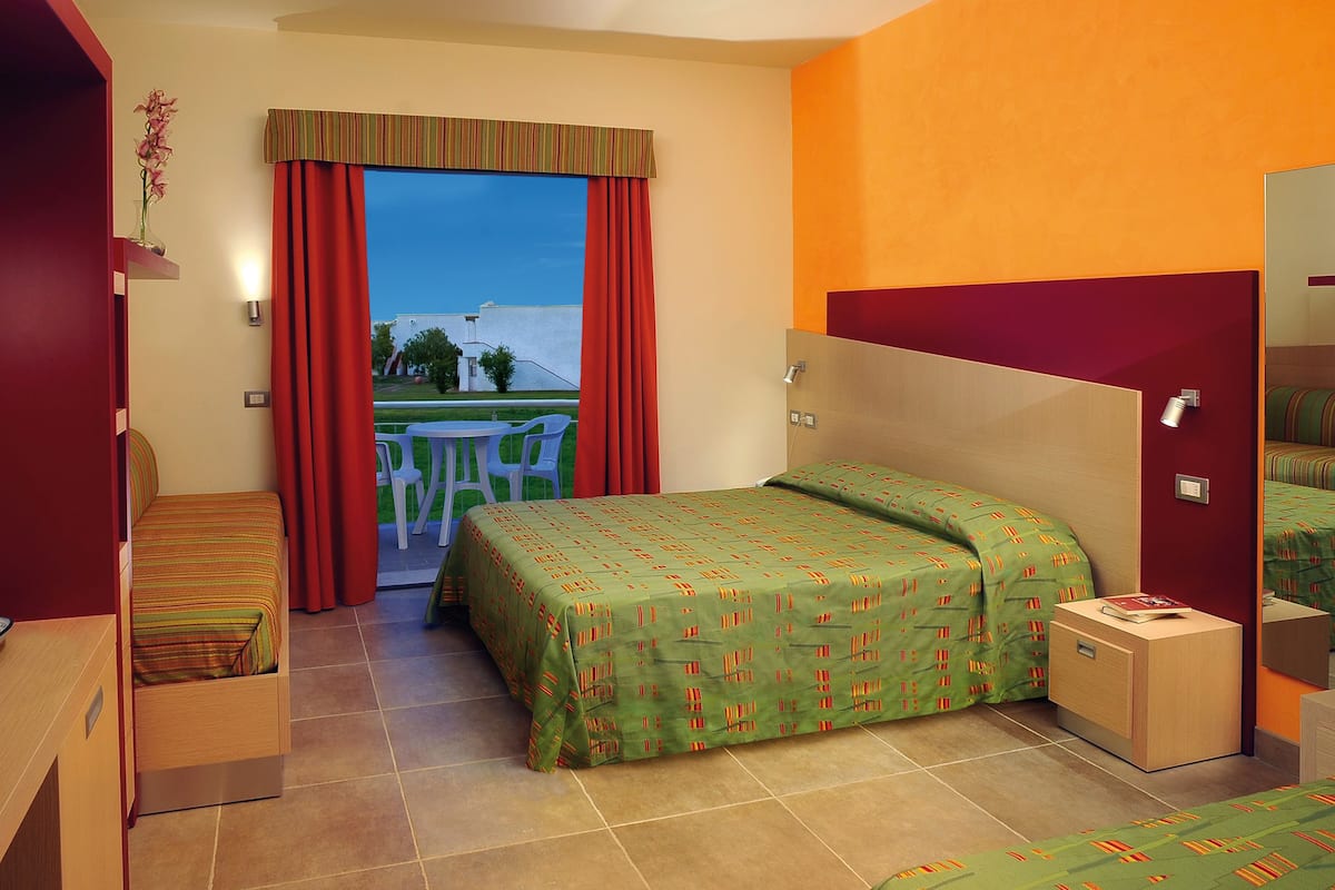 Quarto de hotel com cama, mesa de cabeceira pequena, cadeira, janela com cortinas e vista para o exterior.