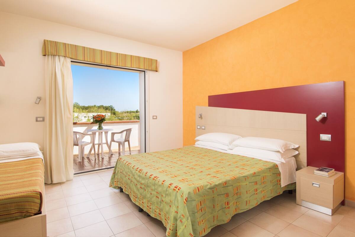 Quarto de hotel com cama, mesa de cabeceira, janela com cortinas, varanda com mesa e cadeiras e vista para o verde lá fora.