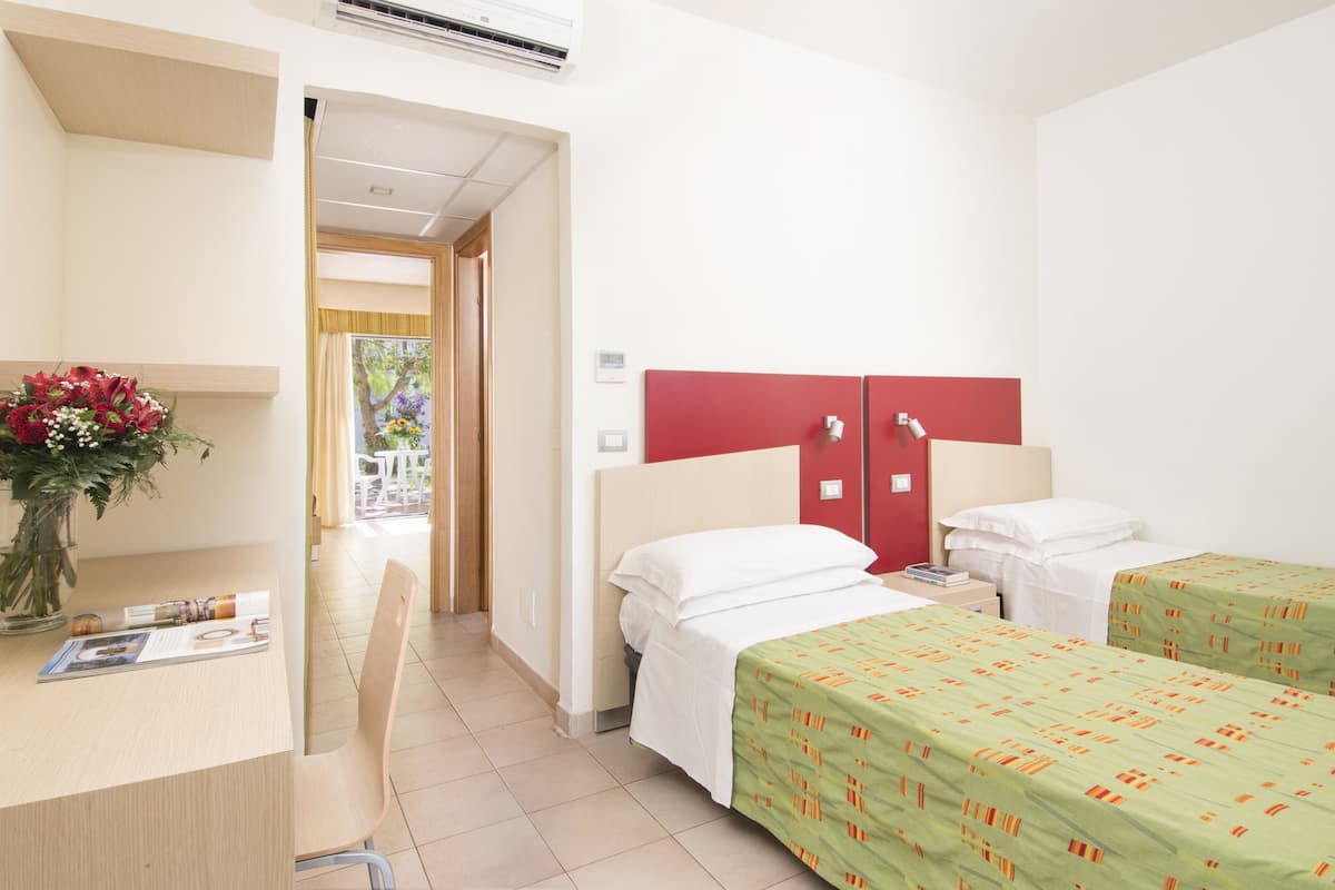 Quarto de hotel com duas camas, uma escrivaninha com cadeira, um guarda-roupa vermelho e vista para o exterior.