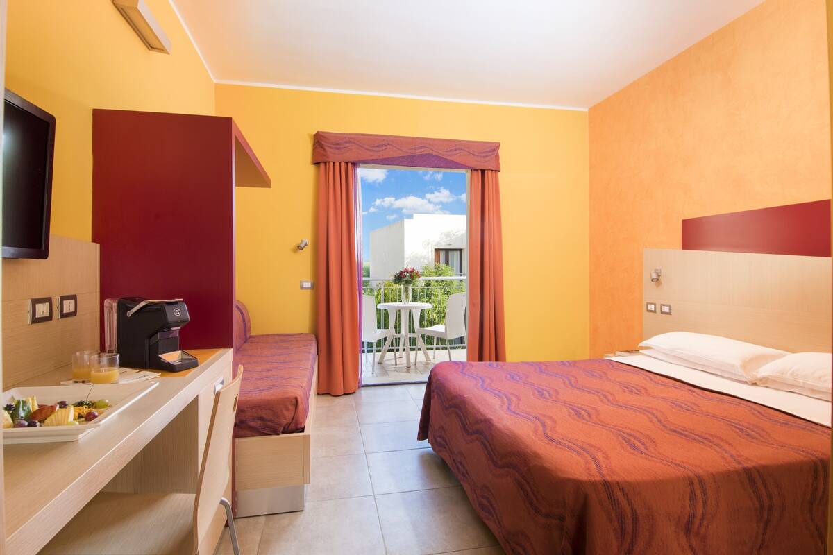 Quarto de hotel com duas camas, uma mesa com cafeteira, televisão e varanda com mesa e cadeiras.