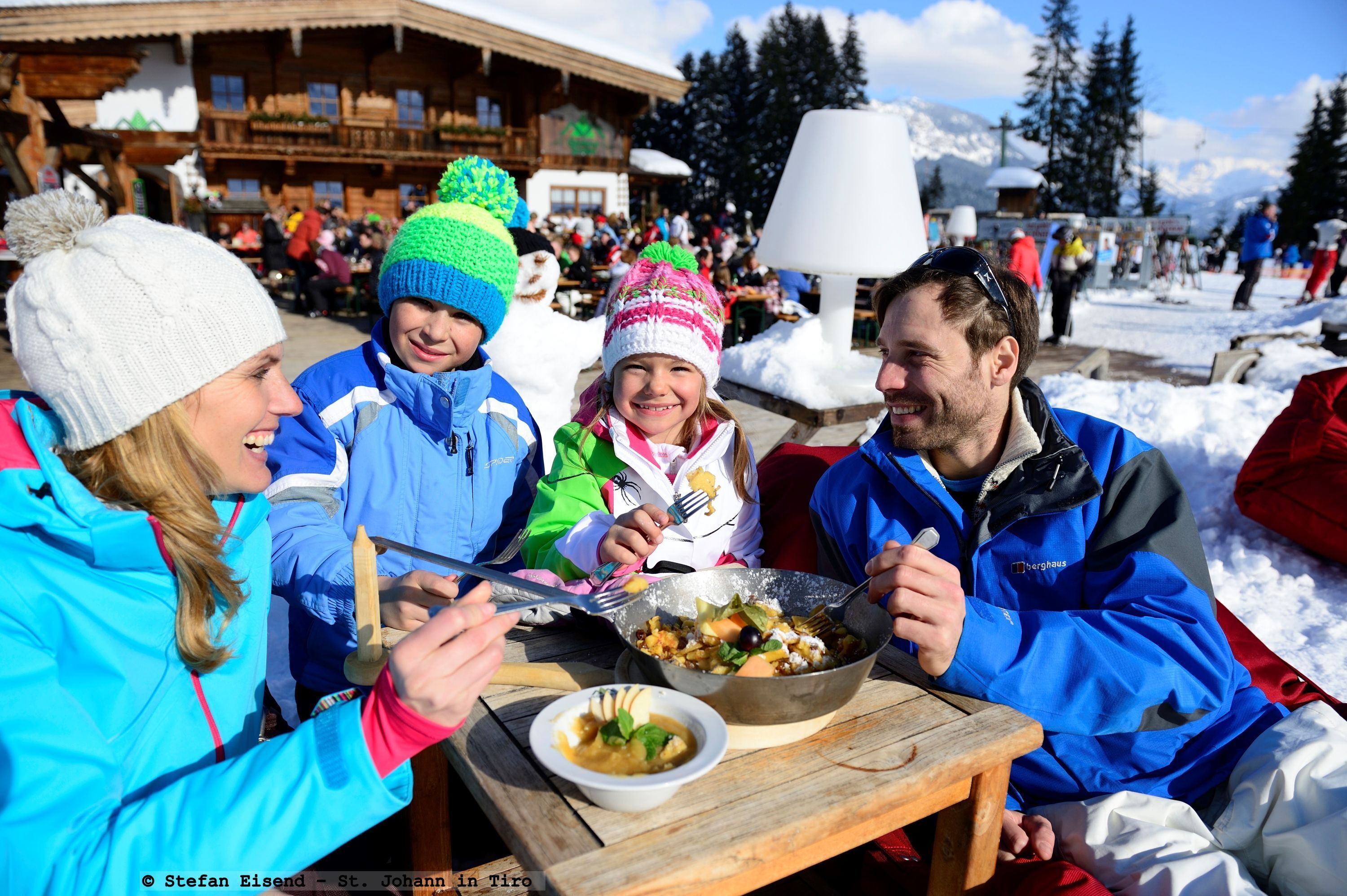 Eine Gruppe von Menschen speist draußen in verschneiter Landschaft, im Hintergrund hölzerne Gebäude.