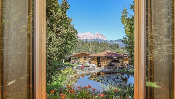 Une cabane en bois avec une cheminée en pierre, un étang parsemé de nénuphars et une vue sur les montagnes en toile de fond.