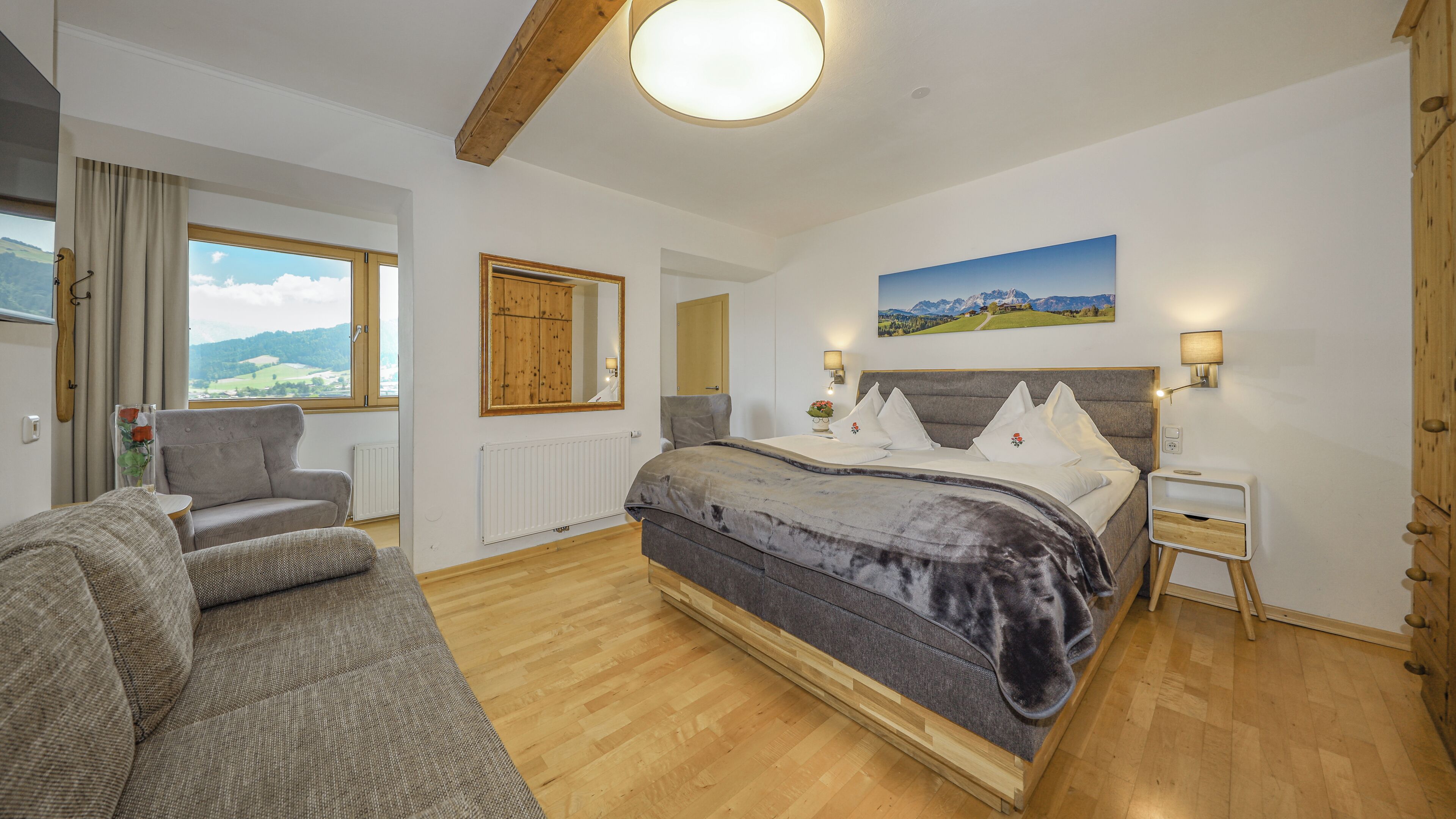 A bedroom with a bed, a sofa, a window with a view, a wooden door, and a painting on the wall.