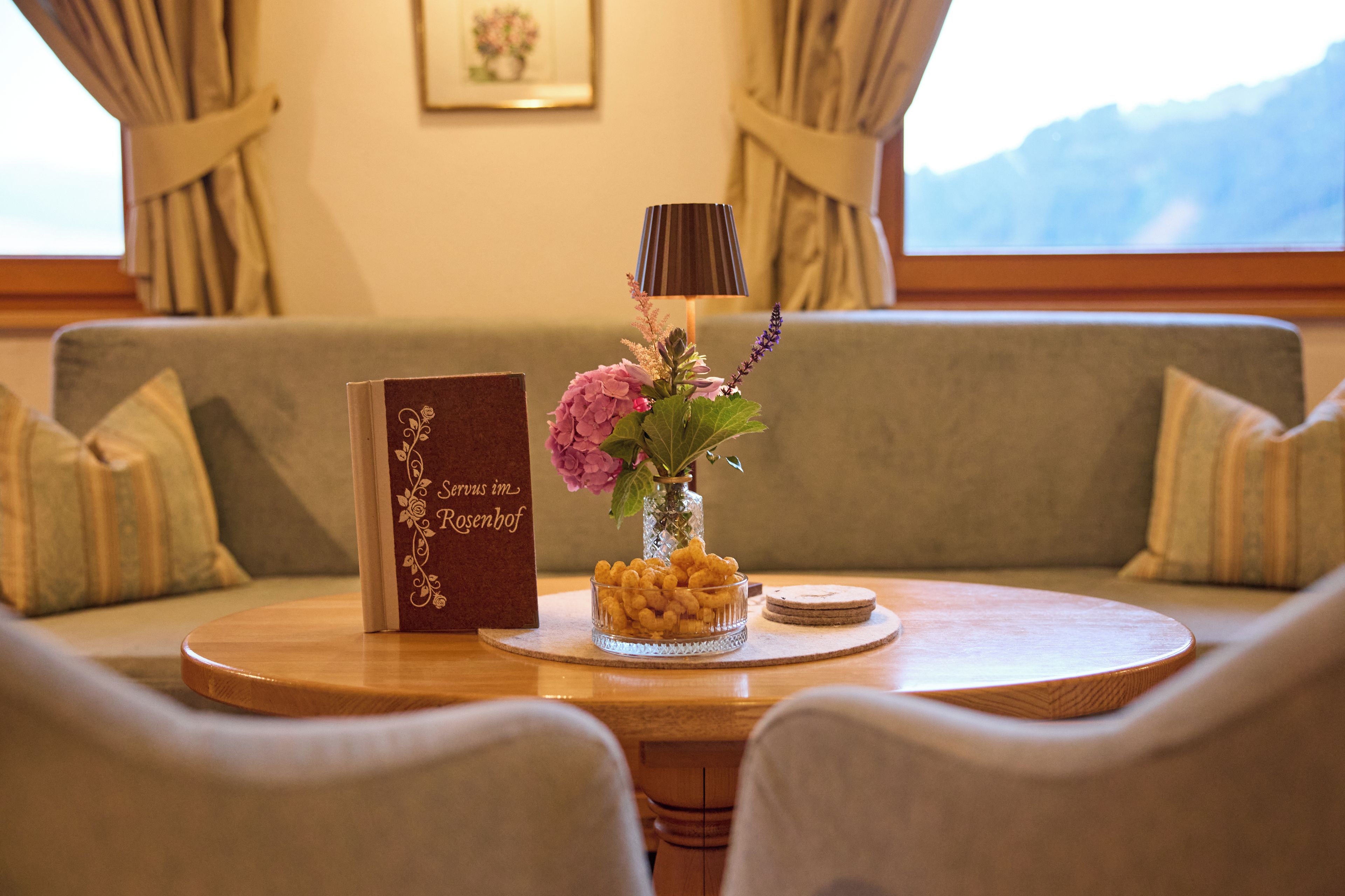 A round wooden table with a book, a vase with flowers, and a glass bowl of snacks.