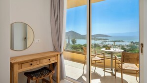 Habitación con un escritorio de madera, un espejo redondo, un taburete pequeño, una mesa redonda con sillas y un amplio ventanal corredizo que da a un balcón con vistas al mar y a las montañas.