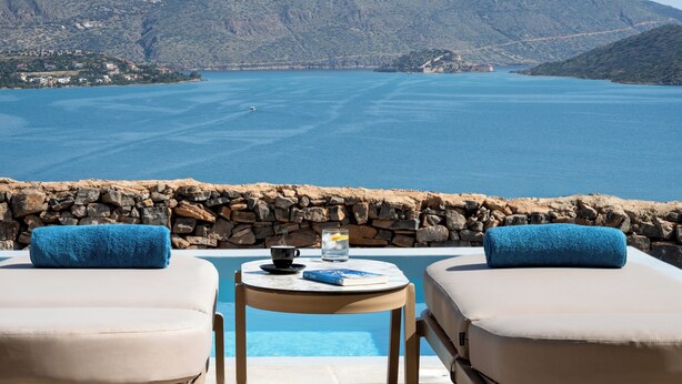 Zona de piscina con dos sillones, una mesa con un libro y una taza, y vistas al mar y colinas.