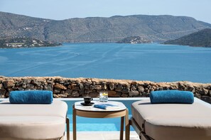 A poolside area with two lounge chairs, a table with a book and a cup, and a view of the sea and hills.