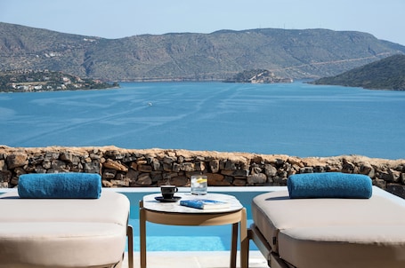 A poolside area with two lounge chairs, a table with a book and a cup, and a view of the sea and hills.