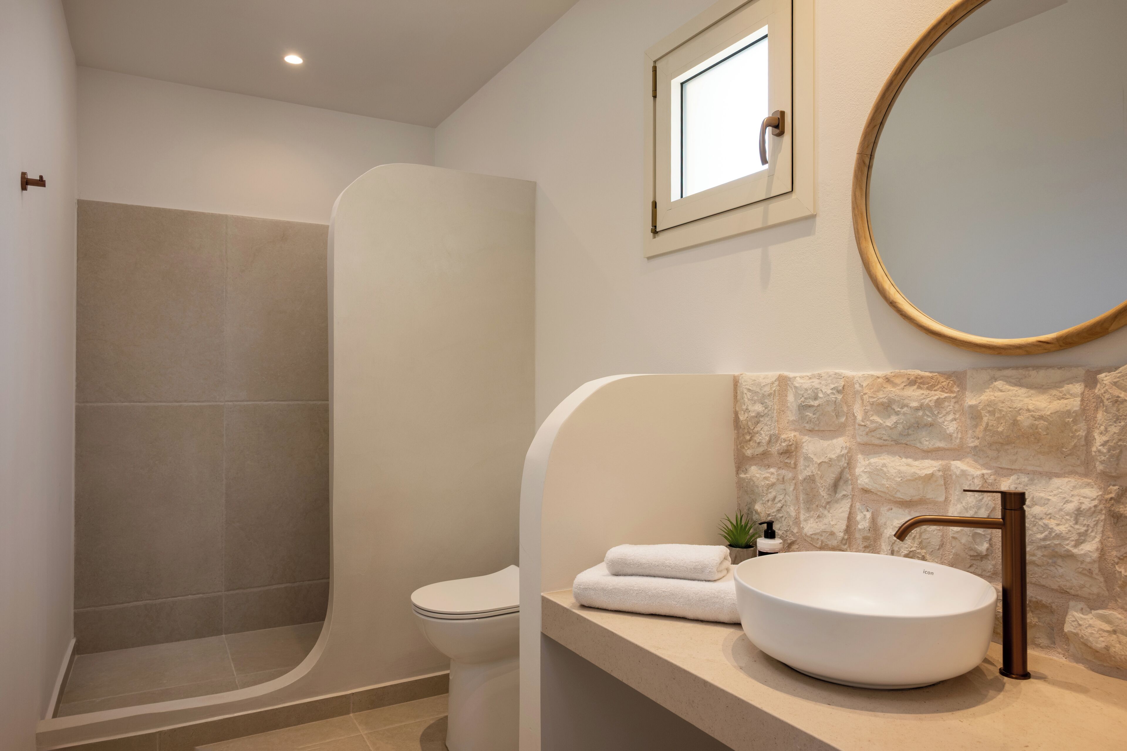 Une salle de bain avec une douche à parois courbes, des toilettes, un lavabo avec un receveur rond et un mur en pierre.