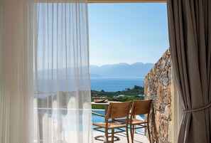 A balcony with a view of the sea and mountains, a table with chairs, and a stone wall. - Elounda Heights - Adults Only (Agios Nikolaos)