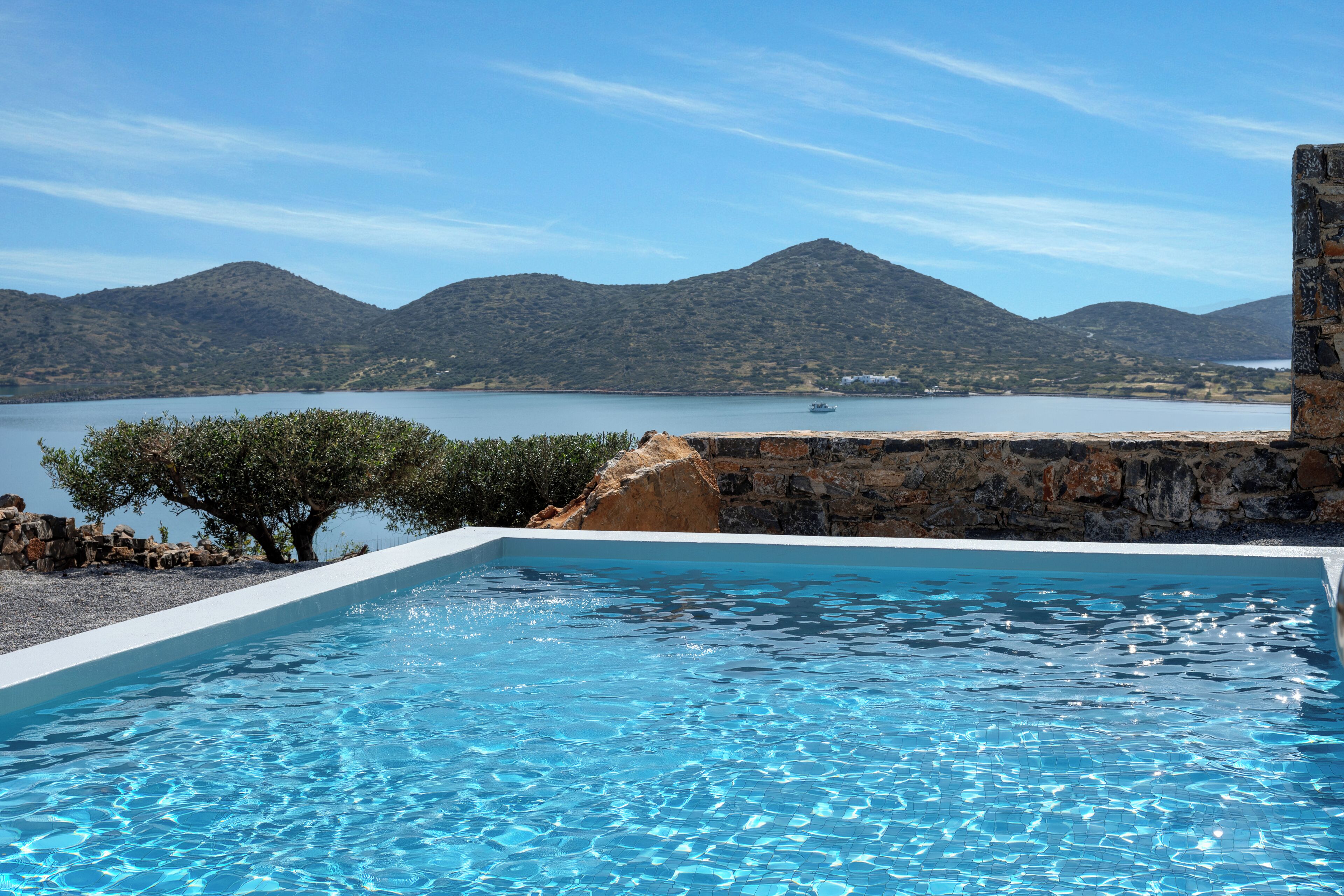 A swimming pool with clear blue water, surrounded by a stone wall and a view of mountains and a lake.