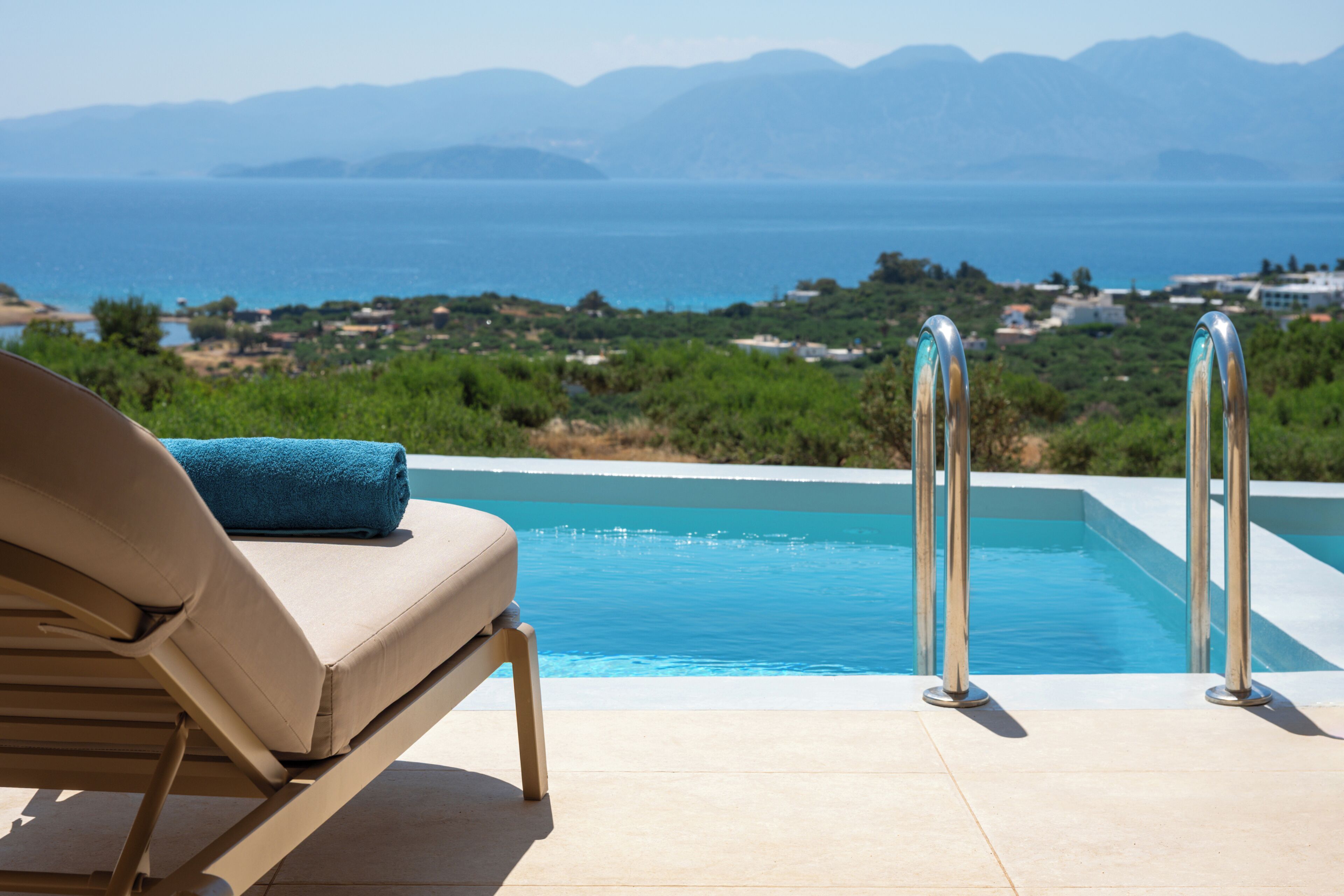Un fauteuil de relaxation placé au bord de la piscine, une serviette à portée de main, une piscine à l’eau d’un bleu limpide et une vue sur la mer et les montagnes lointaines.